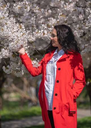 romantisches Bild einer stilvollen Frau in rotem Mantel, in weißer Bluse. Positive Stimmung. Ein nettes Mädchen hält sanft einen Zweig weißen Sakura und blickt lächelnd auf die Blumen.