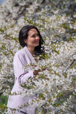 imagen romántica de una mujer con estilo en una chaqueta ligera. Positivo, humor primaveral. Una linda chica suavemente sostiene una rama de sakura blanca y mira las flores, sonriendo.