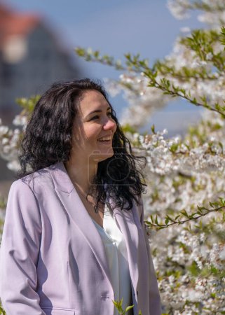 imagen romántica de una mujer con estilo en una chaqueta ligera. Positivo, humor primaveral. Una linda chica suavemente sostiene una rama de sakura blanca y mira las flores, sonriendo.