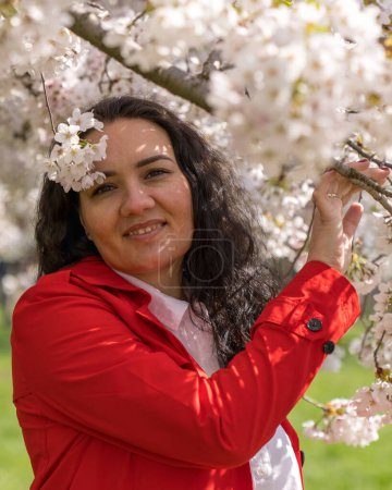 romantic image of a stylish woman in a red coat and white blouse. Positive mood. A cute girl gently holds a white sakura branch and looks at the camera, smiling.