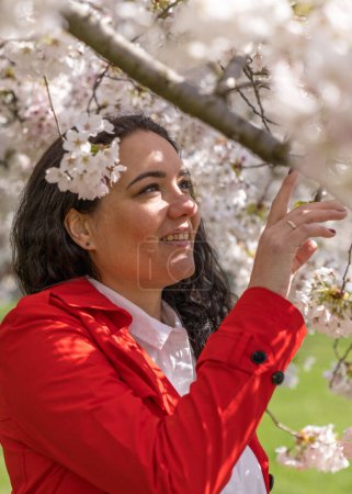 romantic image of a stylish woman in a red coat, in a white blouse. Positive mood. A cute girl gently holds a branch of white sakura and looks at the flowers, smiling