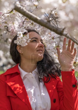 romantic image of a stylish woman in a red coat, in a white blouse. Positive mood. A cute girl gently holds a branch of white sakura and looks at the flowers, smiling