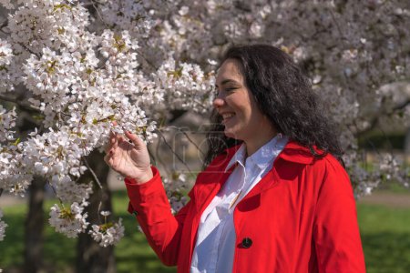 romantisches Bild einer stilvollen Frau in heller Jacke. Positive Frühlingsstimmung. Ein nettes Mädchen hält sanft einen weißen Sakura-Zweig in der Hand und blickt lächelnd auf die Blumen. 