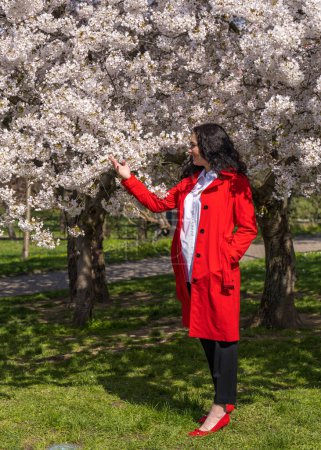romantisches Bild einer stilvollen Frau in heller Jacke. Positive Frühlingsstimmung. Ein nettes Mädchen hält sanft einen weißen Sakura-Zweig in der Hand und blickt lächelnd auf die Blumen. 