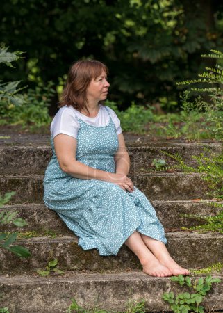 A middle-aged woman sits on old stone steps in an abandoned park. Dressed in a long dress and without shoes, barefoot. Looking thoughtfully into the distance 
