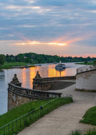 Sonnenuntergang an der Elbe in der berühmten Stadt Dresden. Touristische Ausflugsboote fahren bei Sonnenuntergang langsam den Fluss entlang, vorbei an einer schönen Treppe im Park. Konzept: Urlaub, Tourismus