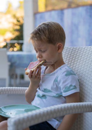 Frühstück vor dem Kindergarten. Netter kleiner Junge isst ein Sandwich mit Wurst, will wirklich schlafen, sitzt mit geschlossenen Augen und isst