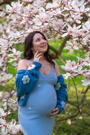 Portrait of a cute pregnant woman in the park, dressed in a blue dress among magnoli flowers. The expectant mother looks tenderly at a flowering tree. Pregnancy. Blooming magnolia 