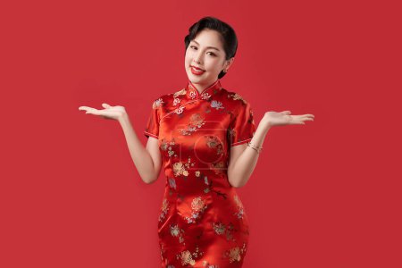 Téléchargez les photos : Bonne année chinoise, Belle jeune femme asiatique portant la robe traditionnelle cheongsam qipao avec geste d'introduction isolé sur fond rouge, - en image libre de droit