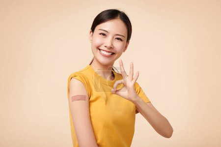 Saludable mujer asiática recibiendo inmunidad vacunada dando señal de mano ok en fondo beige, concepto de inoculación recomendada, vacunación, 