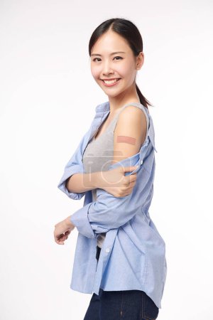 Photo for Young asian women smiling after getting a vaccine, holding down her shirt sleeve and showing her arm with bandage after receiving vaccination on white background, - Royalty Free Image