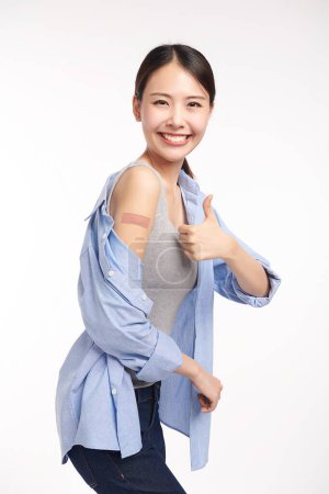 Photo for Young asian women smiling after getting a vaccine, holding down her shirt sleeve and showing her arm with bandage after receiving vaccination on white background, - Royalty Free Image