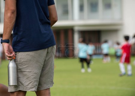 Dad standing and watching his son playing football in a school tournament on a sideline with a sunny day. Sport, outdoor active, lifestyle, happy family and soccer mom and soccer dad concept.