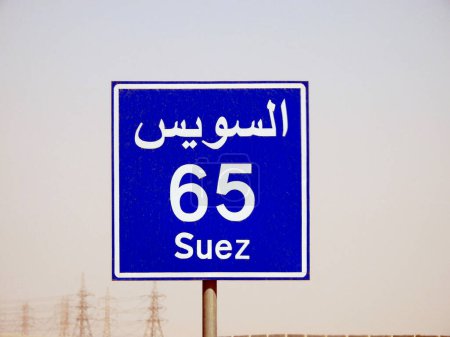 Foto de Un letrero de carretera en la carretera de Suez El Cairo da la distancia restante a la ciudad de Suez 65 KM sesenta y cinco kilómetros escritos en Inglés y la palabra Suez escrito en árabe e inglés al lado de la calle - Imagen libre de derechos