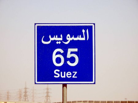 Foto de Un letrero de carretera en la carretera de Suez El Cairo da la distancia restante a la ciudad de Suez 65 KM sesenta y cinco kilómetros escritos en Inglés y la palabra Suez escrito en árabe e inglés al lado de la calle - Imagen libre de derechos