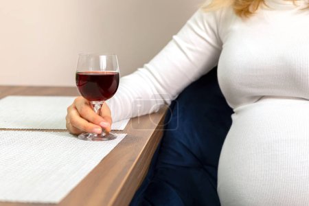 Foto de Cultivado blanco caucásico hembra embarazada sostiene, bebe un vaso de vino tinto sin alcohol, sentado en la silla en la cocina. Bebida no alcohólica. La mujer lleva una manga larga blanca. Relajación por la noche - Imagen libre de derechos