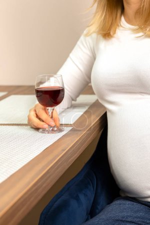 Foto de Cultivado blanco caucásico hembra embarazada sostiene, bebe un vaso de vino tinto sin alcohol, sentado en la silla en la cocina. Bebida no alcohólica. La mujer lleva una manga larga blanca. Relajación por la noche - Imagen libre de derechos