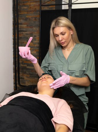 Photo for Acne treatment, young skin care. Cosmetologist does alginate mask procedure on female teen face. Teenage girl lying on medical couch. Dermatologist in pink gloves holds bowl, brush. Vertical plane. - Royalty Free Image