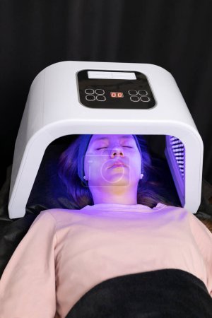 Cosmetic led light face mask. Young woman, teenage girl lying under facial regenerative treatment mask. Health and beauty face skin care. Acne, beauty spot cure. Beauty Photon Therapy. Vertical plane