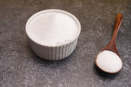 Erythritol, organic sweetener, produced by fermentation from corn, called dextrose in ceramic bowl, wooden spoon on granite background, table. Sugar substitute. Horizontal plane.