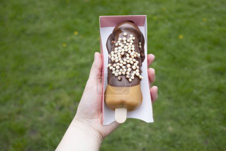 Photo for Festival Street Food Penis Shaped Waffle On Wooden Stick With Dark Chocolate, Decorated With White Air Corn Balls In Paper Box In Human Hand Outdoor. Sweet Cookie Horizontal Plane. - Royalty Free Image
