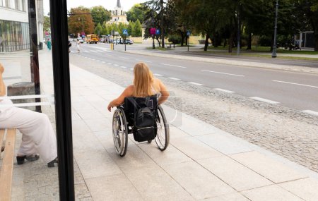 Foto de Vista trasera Adulto con baja estatura en silla de ruedas se para en la parada de autobús al aire libre esperando el transporte público en el día de verano. Mujer adulta con discapacidad. Copiar espacio para texto. Transporte. Horizontal. - Imagen libre de derechos