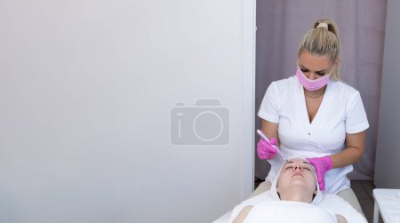 Foto de Cosmetólogo de la bandera que hace la inyección de la mesoterapia con Dermapen en la cara de la mujer joven hermosa para el rejuvenecimiento en salón del balneario. Paciente recibiendo mesoterapia con aguja, cuidado de la piel. Espacio de copia horizontal. - Imagen libre de derechos