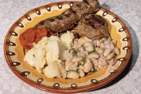 Photo for Closeup Plate With Traditional Bulgarian Food. Mashed Potato, White Beans, Haricot, Vegetable Relish Ljutenica, Grilled Minced Meat With Spices Kebapche. Horizontal Plane. High Quality Photo - Royalty Free Image