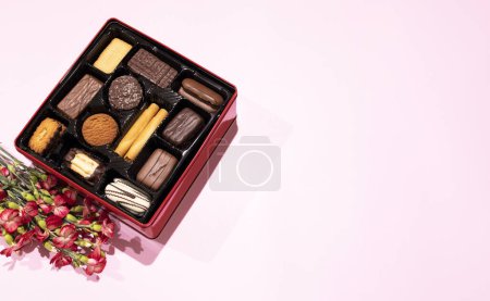 Verschiedene Shortbread Belgian Cookie In Red Box auf rosa Hintergrund mit frischen Blumen. Festliches Dessert-Sortiment für Feiertage. Flache Lay 12 Abschnitte Feature Leckereien, Kekse horizontal, Kopierraum