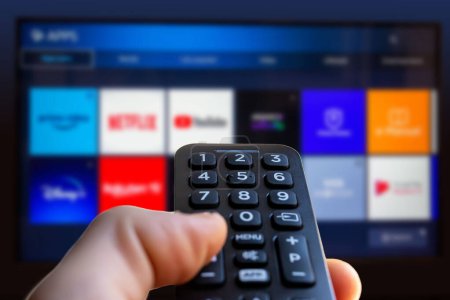 A man is holding the remote control of a smart TV with a television screen in the background with some blurry video streaming service app icon