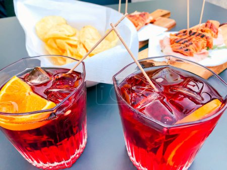 Téléchargez les photos : Cocktail rouge avec glace, tranche d'orange et collation salée aux chips. Cocktail frais, savoureux et coloré sur une table. - en image libre de droit