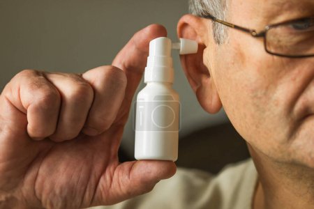 Hombre mayor caucásico en gafas utiliza un spray para los oídos. La mano de Mans sostiene una botella de pulverización de oído blanco con boquilla instalada en el orificio de la oreja. Higiene diaria y limpie los oídos de la cera. Concepto de salud.