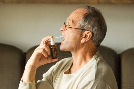 Photo for Sick caucasian senior man with open mouth using a throat spray against a dry cough. Side view. Old man with glasses in white warm sweater treates at home. Seasonal diseases. Health care concept. - Royalty Free Image