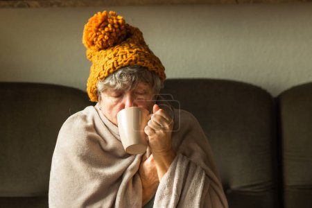 Téléchargez les photos : Femme âgée en bonnet de gingembre tricoté enveloppé dans une couverture et boire une tasse de thé chaud. Vieille grand-mère se sent froid à la maison avec des problèmes de chauffage. La pauvreté et la crise énergétique en Europe. Symptômes de froid. - en image libre de droit