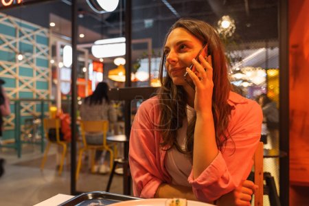 Kaukasische brünette Frau, die im Café am Smartphone spricht. Eine Frau in lässiger Kleidung hält ihr Handy in der Hand, während sie in einem chinesischen Lokal auf ihre Bestellung wartet. Menschen und Gadgets-Konzept