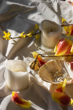 Photo for Morning still life with candles, vintage bell extinguishing, red and yellow tulip petals, jewelry: earrings, pearls in seashell on white sheet of bed. Female spring background. Women's Day concept. - Royalty Free Image