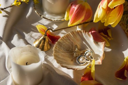 Photo for Morning still life with candles, vintage bell extinguishing, red and yellow tulip petals, jewelry: earrings, pearls in seashell on white sheet of bed. Female spring background. Womens Day concept. - Royalty Free Image