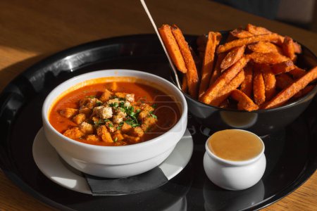 Photo for Bowl of red tomato soup gazpacho or borscht with croutons, vegetables and herbs, bowl of sweet potato fries with sweet chili sauce on black tray in cafe. Lunch break concept. Photo for restaurant menu - Royalty Free Image
