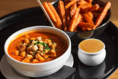 Photo for Bowl of red tomato soup gazpacho or borscht with croutons, vegetables and herbs, bowl of sweet potato fries with sweet chili sauce on black tray in cafe. Lunch break concept. Photo for restaurant menu - Royalty Free Image