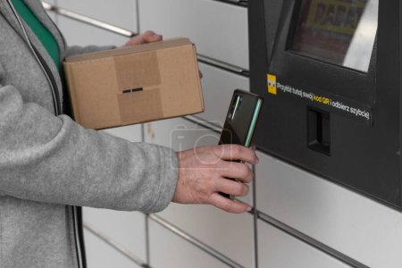 Pniewy, Poland - 22 March 2023. Senior woman with smartphone entering QR code to open shopping locker in inPost delivery service and send parcel. E-commerce shipping and delivery. People and gadgets.