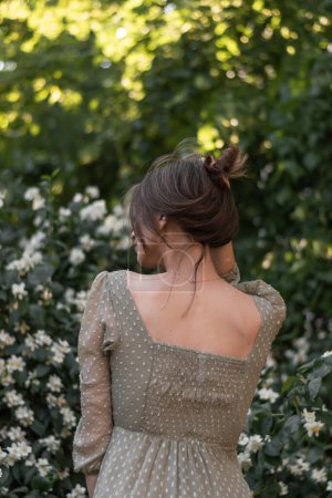 Photo for Beautiful young woman in vintage dress stands with her back and gathered her hair in a bun. Flowering jasmine tree background. Female enjoys the fragrance of blooming bush, tranquil scene. Back view. - Royalty Free Image