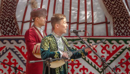 Téléchargez les photos : Almaty, Kazakhstan - 18 septembre 2022. Les jeunes hommes en costumes nationaux kazakhs chantent et jouent de la dombra instrument de musique folklorique sur une scène de rue stylisée comme une yourte. Concours de chanteurs - aitys - en image libre de droit