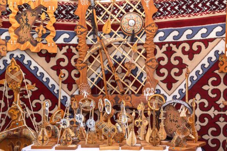 Téléchargez les photos : Souvenirs faits à la main dans le style kazakh - modèles d'instruments folkloriques de musique, armes de guerriers anciens, décorations murales en bois et en cuir sur le fond des murs en feutre de la yourte avec des ornements asiatiques traditionnels. - en image libre de droit