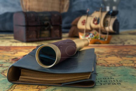 Photo for Spyglass and a leather-bound book lie on an old map against the background of a forged chest and a wooden model of a sailboat. Close-up - Royalty Free Image