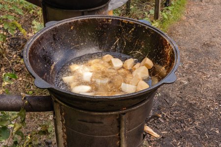 Photo for Coarsely chopped potatoes are fried in boiling oil in a cast-iron cauldron on a wood-burning stove. Street food - Royalty Free Image