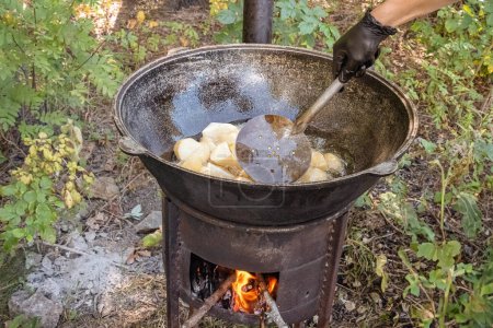 Photo for Cook uses a slotted spoon to mix coarsely chopped potatoes in boiling oil in a cast-iron cauldron on a wood-burning stove. Street food - Royalty Free Image