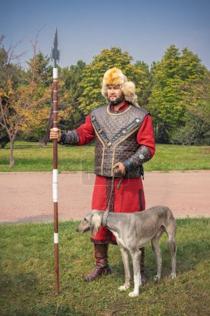 Téléchargez les photos : Almaty, Kazakhstan - 17 septembre 2023. Jeune homme en costume national d'un guerrier kazakh se tient avec une lance de bataille et un chien de chasse de la race Tazy. Portrait - en image libre de droit