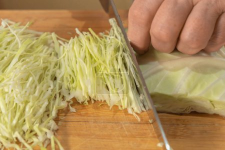 A Cook thinly slices white cabbage with a knife on a wooden cutting board to prepare a vegetable salad. Close-up. Veganism and raw food diet concept.