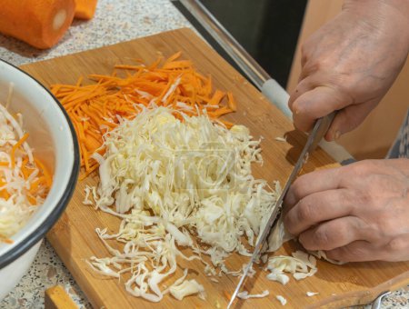 The cook thinly cuts white cabbage with a knife on a wooden cutting board to prepare a vegetable salad with carrots. Veganism and raw food diet concept.