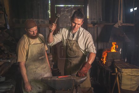 El aprendiz de herrero forja una pieza de metal al rojo vivo en un yunque en una forja bajo la supervisión de un maestro. Formación en artesanía